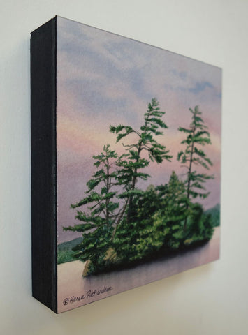 A watercolor painting of a small island with tall green pine trees under a soft pink, purple, and blue sky, with calm water in the foreground. Titled Evening Magic by Karen Richardson