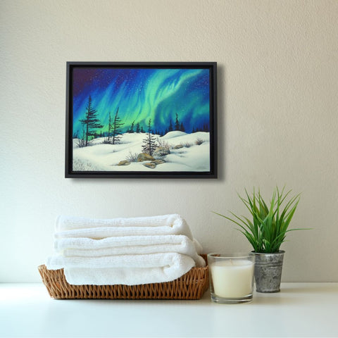Framed artwork of a snowy landscape with trees and a starry sky, titled Dancing With the Stars, by Karen Richardson, placed above a stack of towels in a bathroom setting.