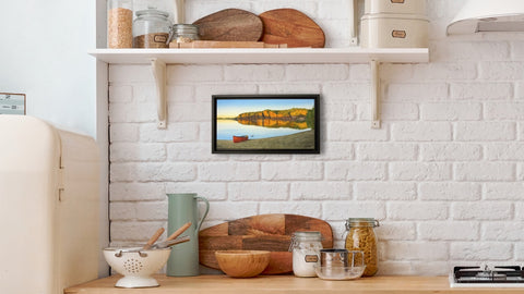 Small framed picture of a red canoe on a beach with a scenic landscape of cliffs and water, displayed on a white brick wall above a wooden shelf with kitchen items. Painting titled Mazinaw Mirror by  Karen Richardson