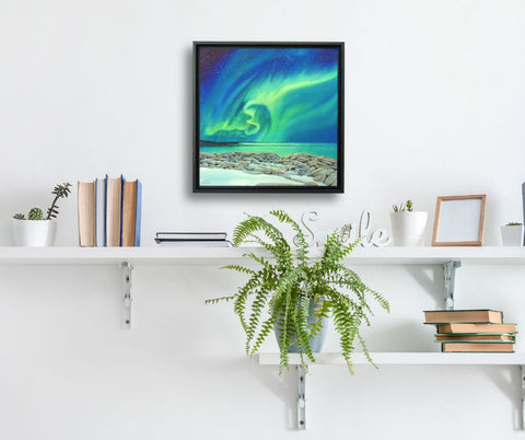 Framed artwork of the Northern Lights titled Rhythms of the Night, by Karen Richardson, shown on a white wall above a shelf with books and plants.