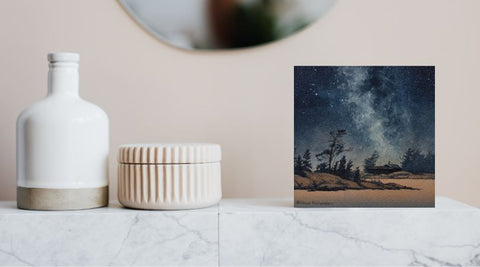 Painting of a starry night sky over a silhouette of trees and a cabin, titled Finding Peace, by Karen Richardson, shown in a bathroom setting.